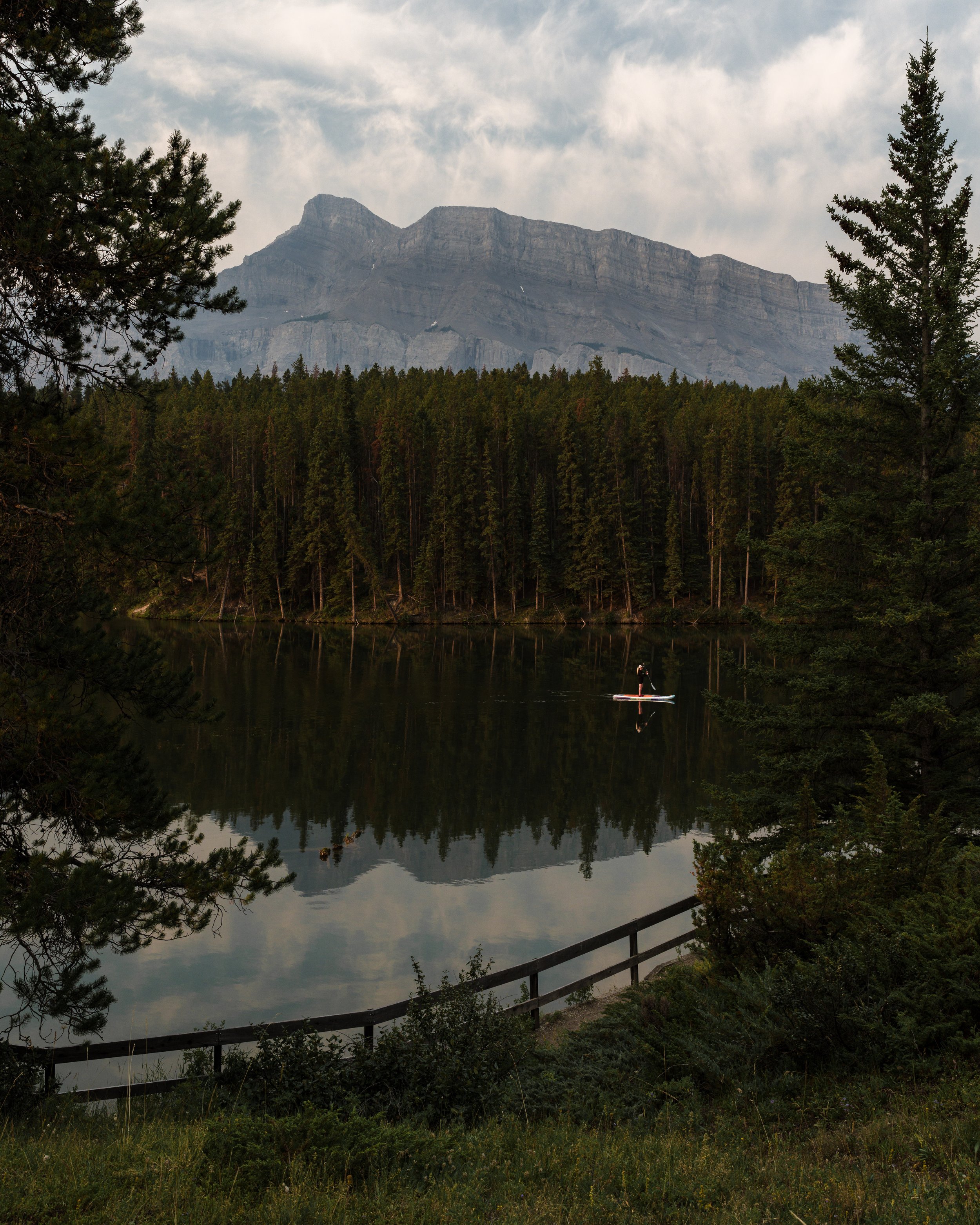 A serene lake surrounded by dense evergreen forest with mountain peaks in the background and a person stand-up paddleboarding on the water.