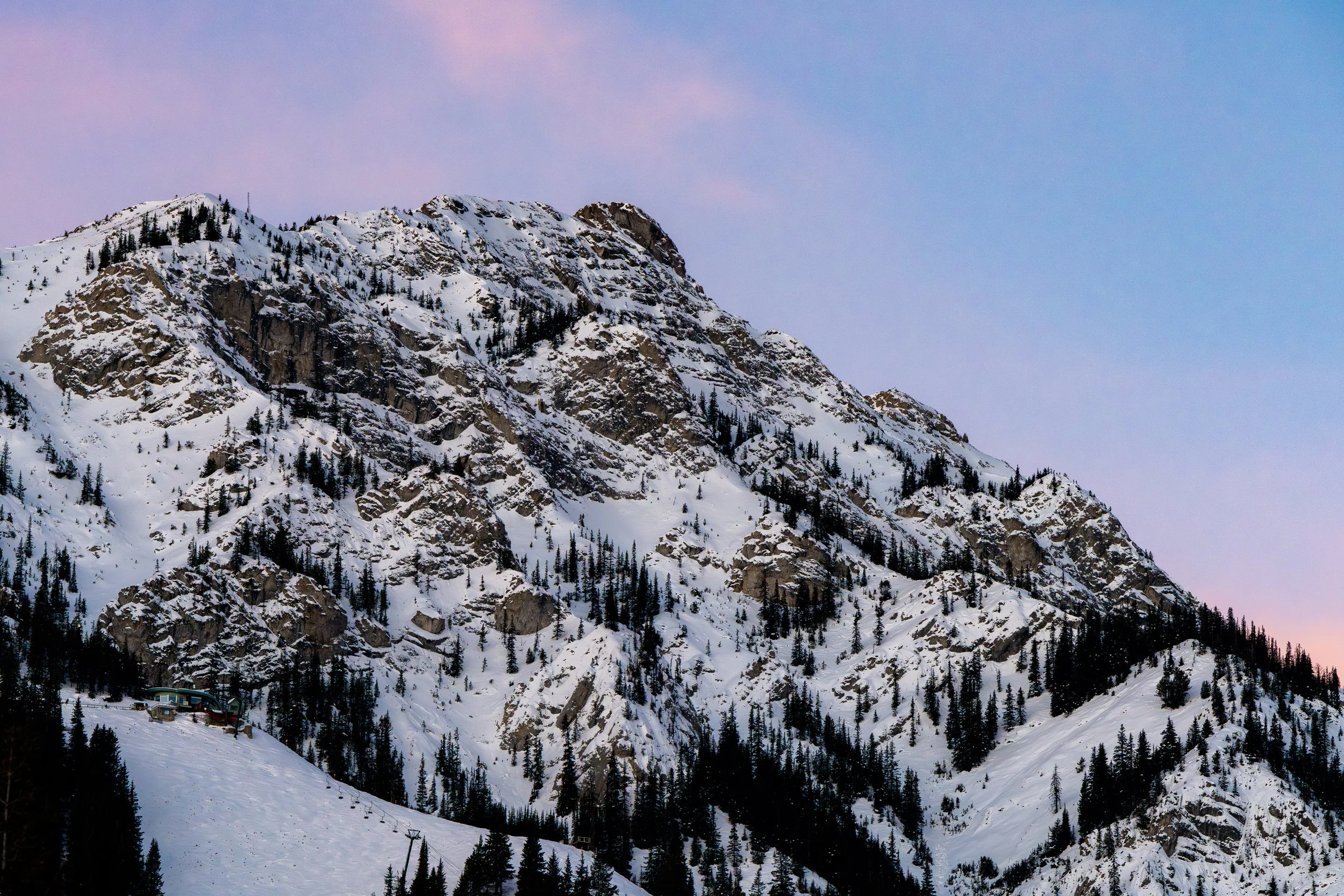 Snow-capped mountain with evergreen trees and a small building at the base, under a partly cloudy sky.