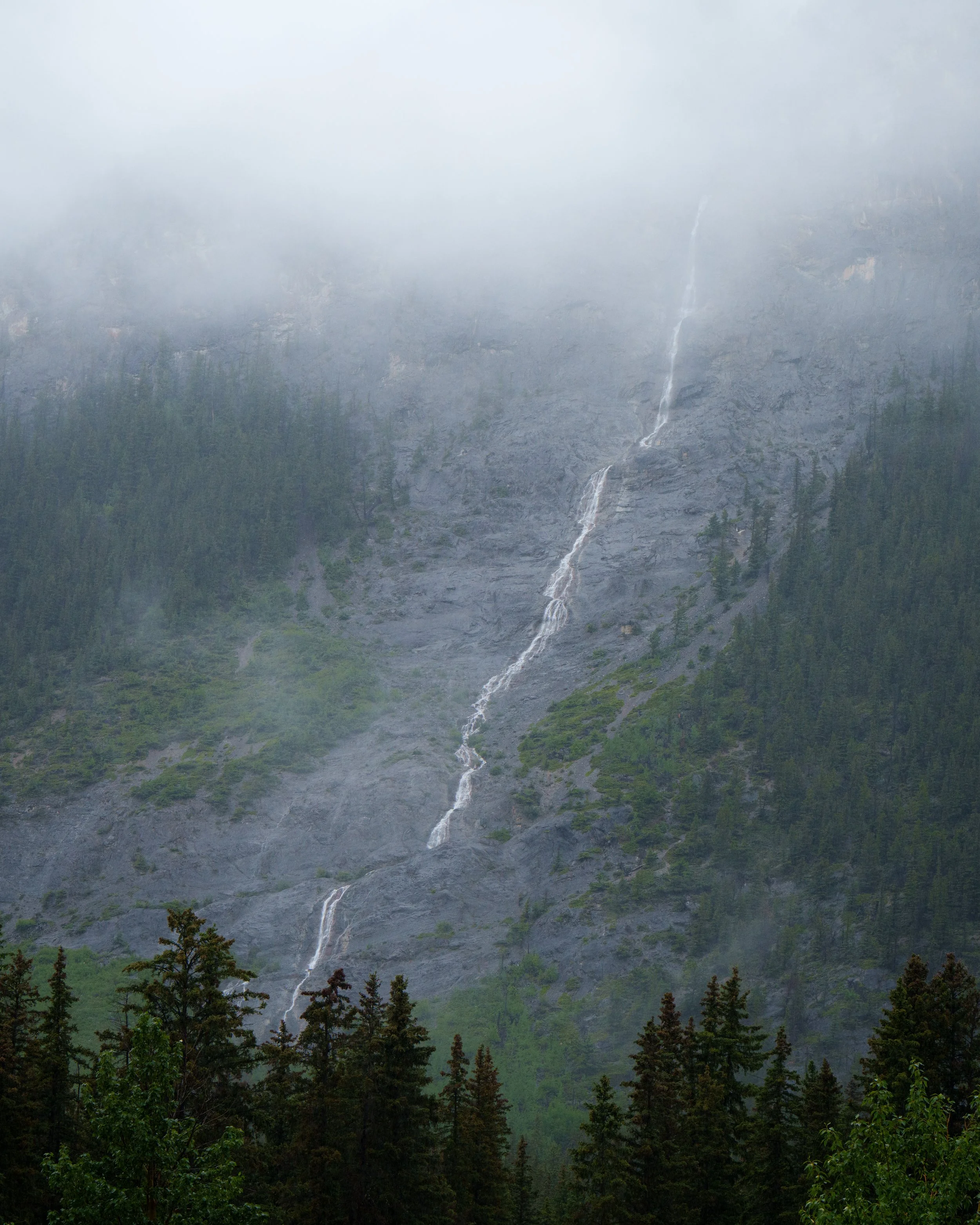A mountain with a waterfall cascading down its rocky face, partially shrouded in fog, with trees at the bottom.