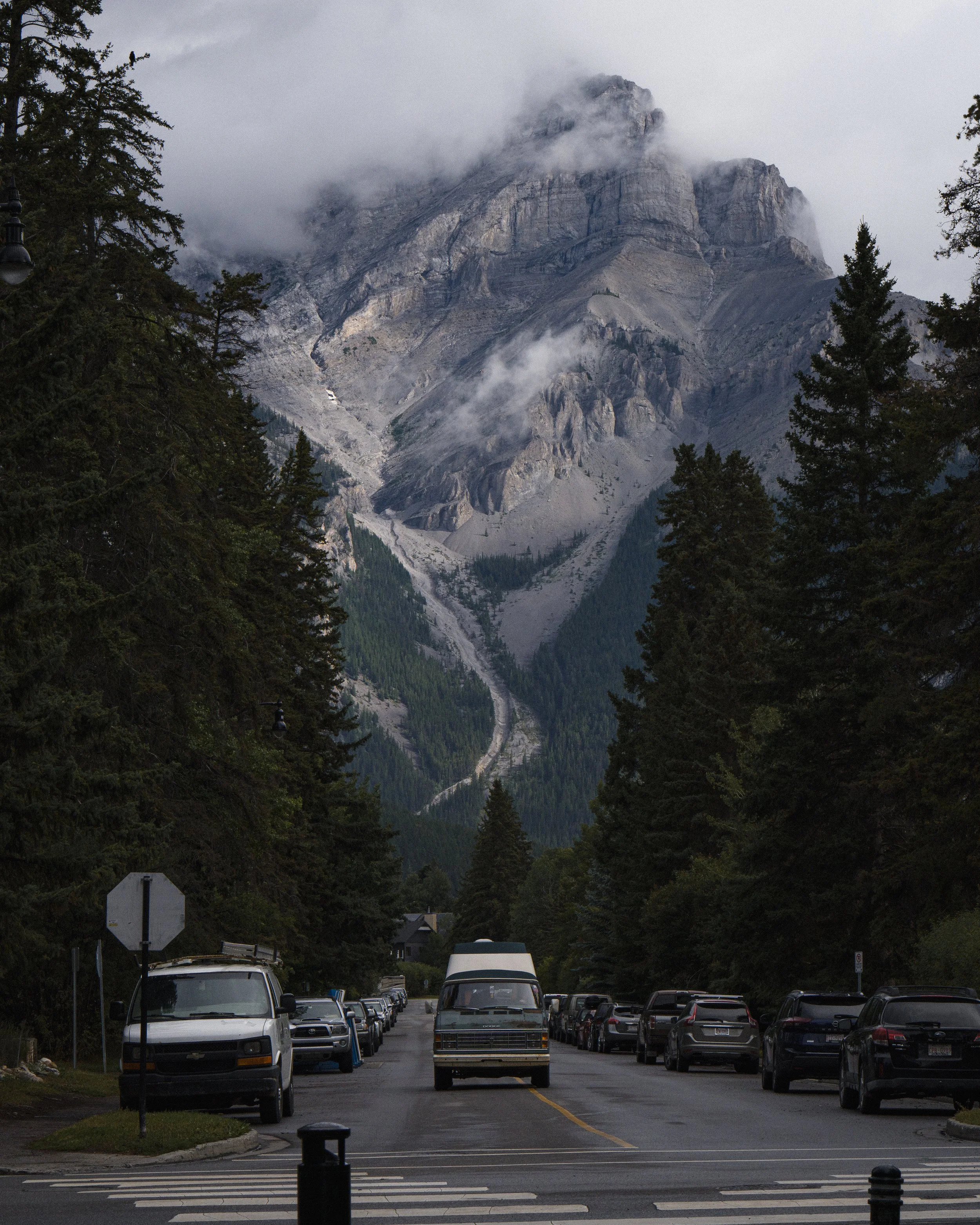 A mountain landscape with a glacier and clouds, viewed from a parking lot lined with cars and surrounded by tall pine trees.
