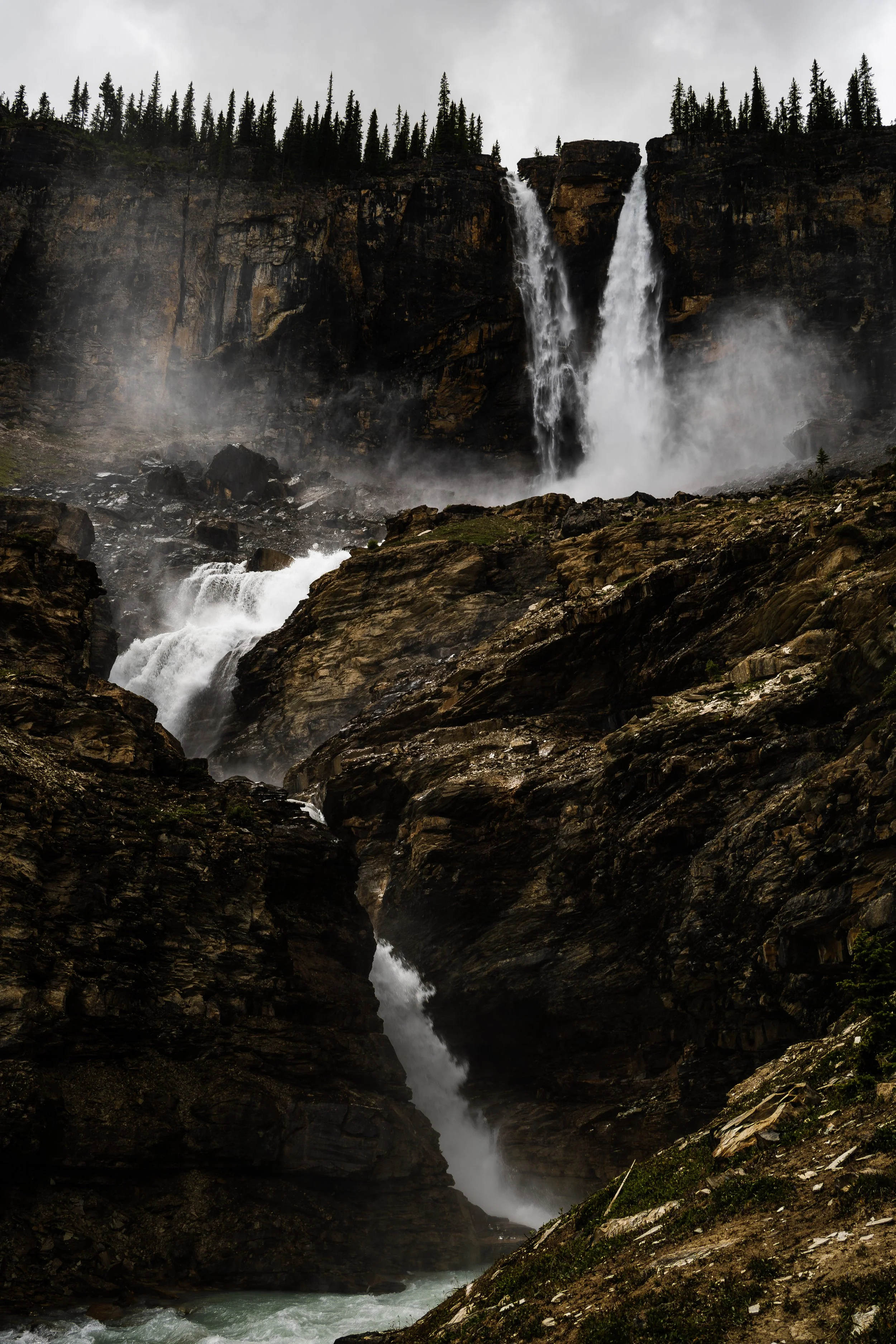 Multiple waterfalls cascading down a rocky cliffside surrounded by pine trees and misty skies.