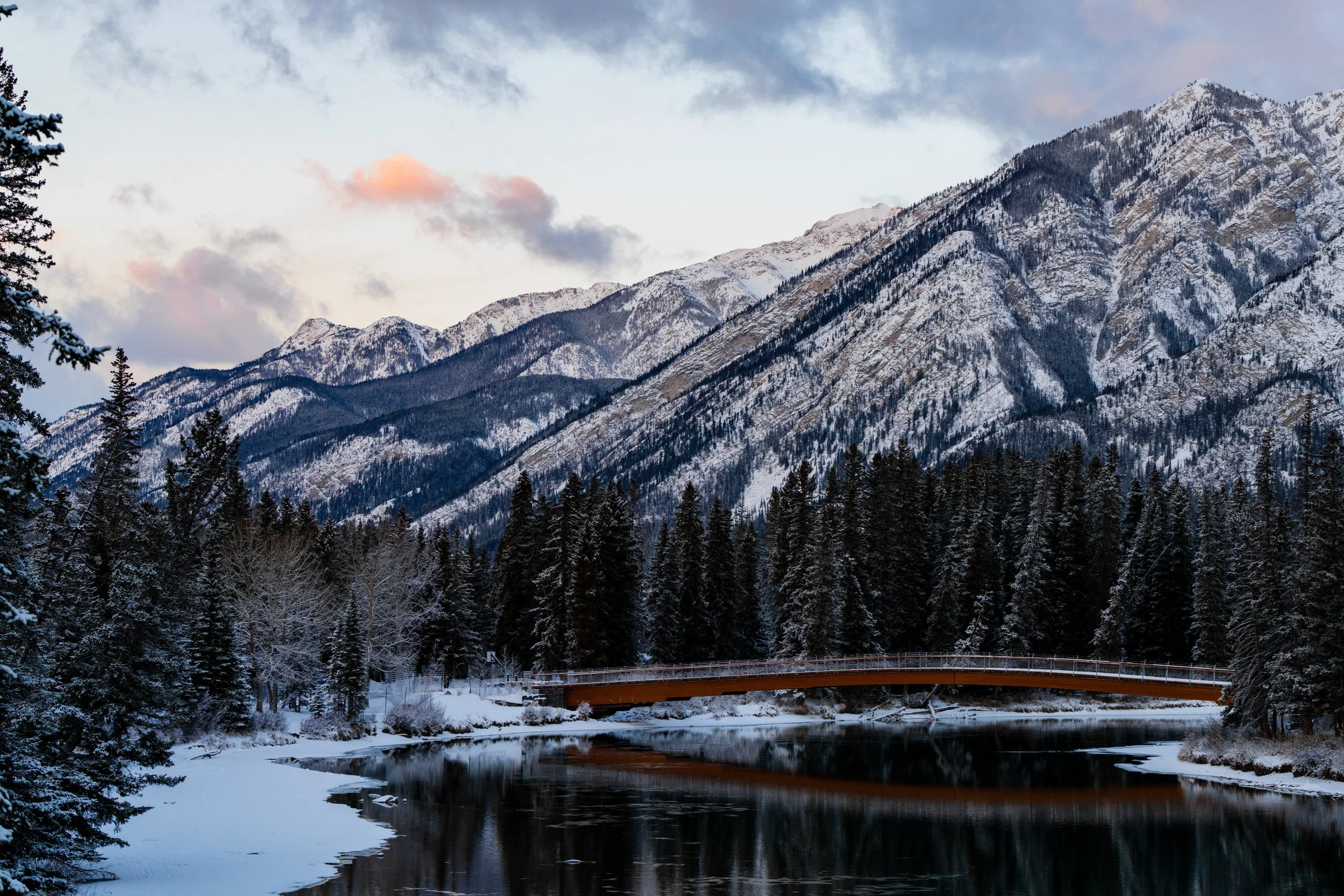 Snow-covered mountains and dense pine forest beside a partially frozen river with a wooden bridge