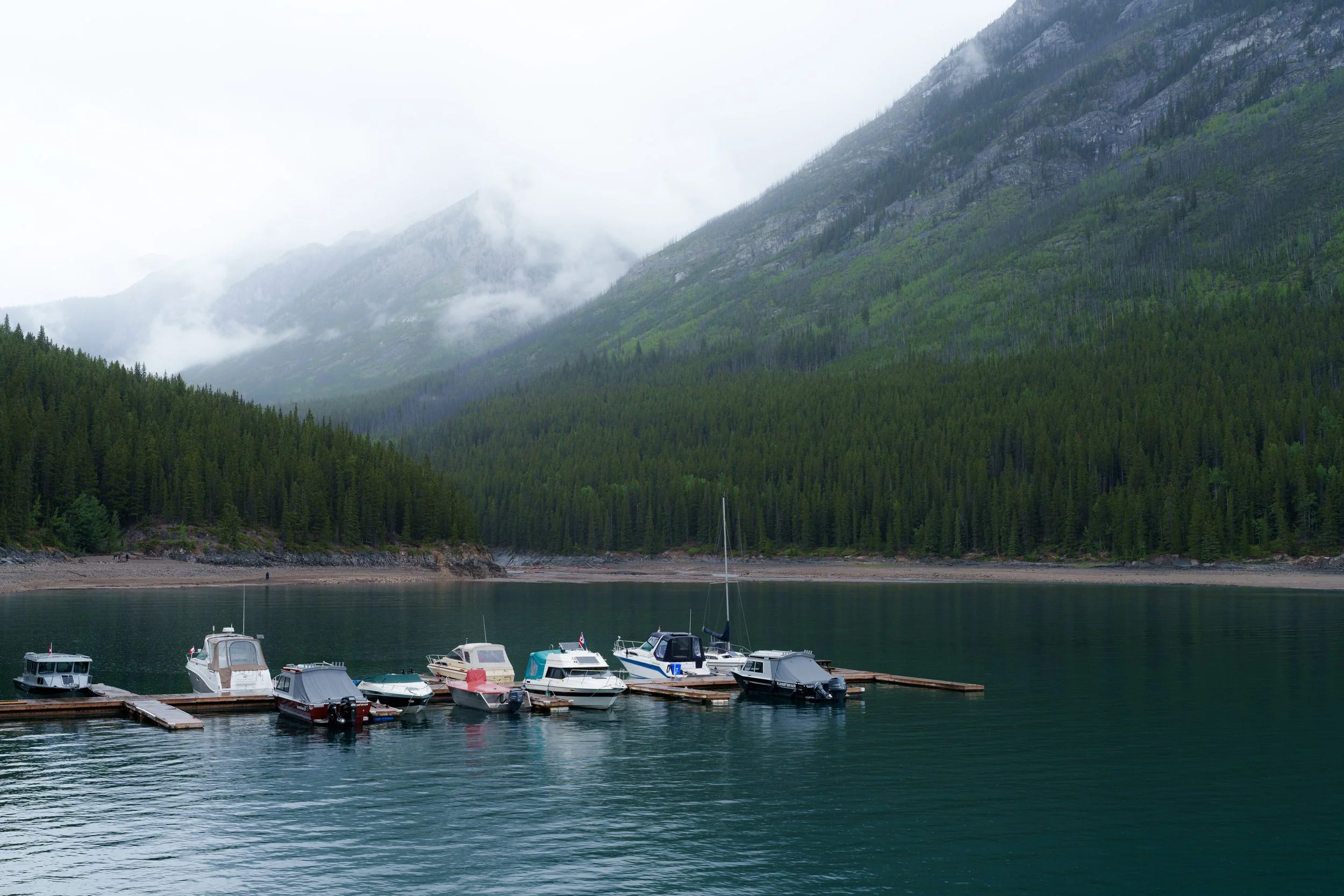 Boats docked at a marina on a calm lake with evergreen trees and mountains in the background, shrouded in mist.