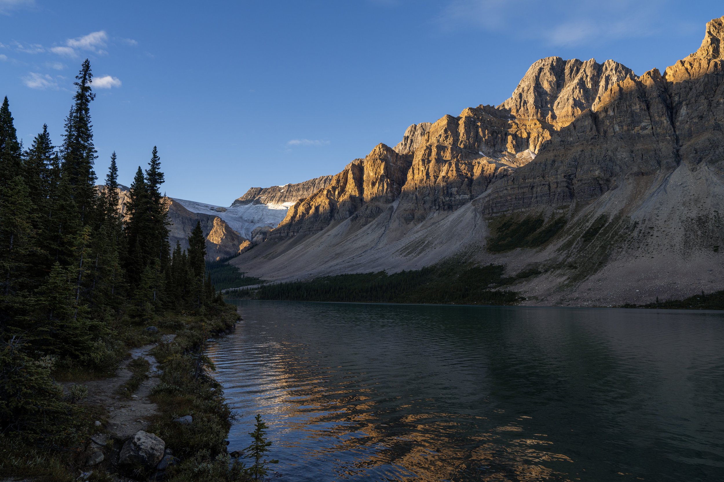 A mountain lake with a forested shoreline and towering rocky mountains in the background, sunlight illuminating the peaks.