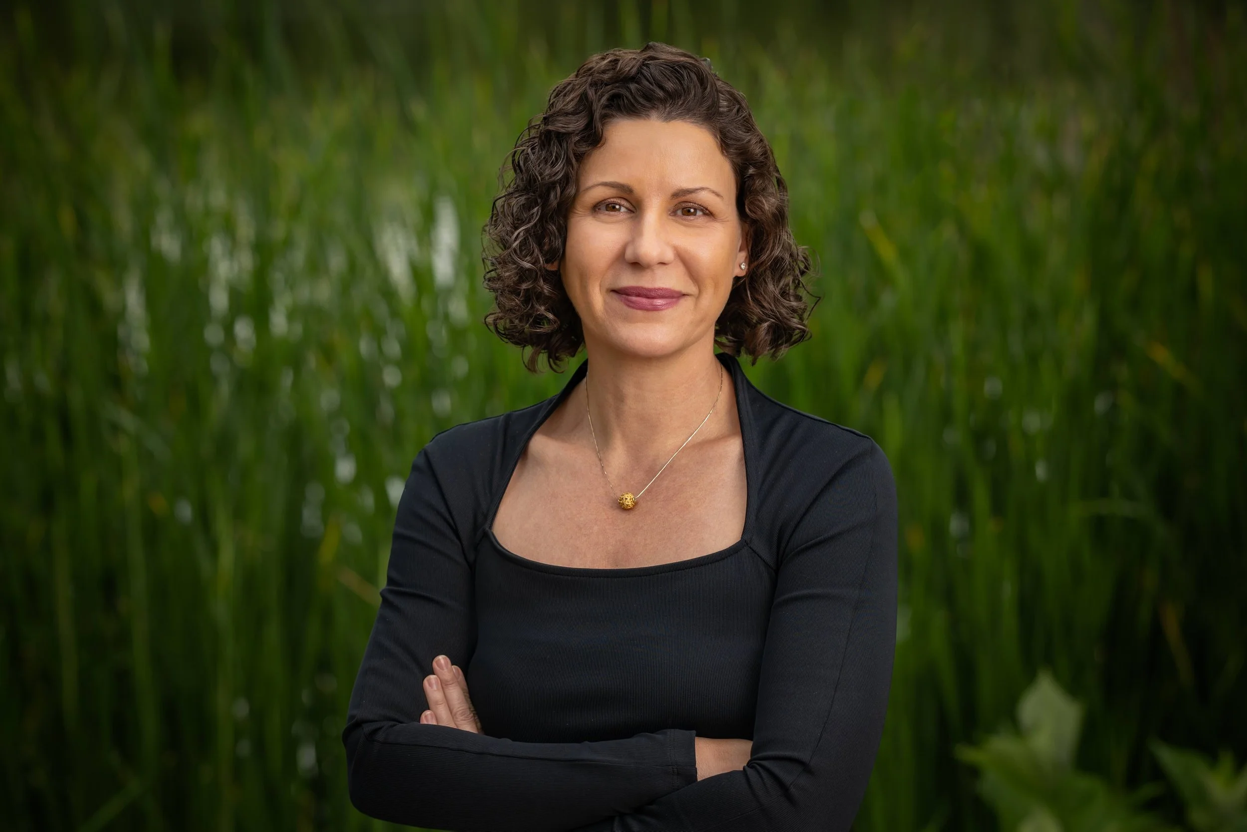 A woman with curly brown hair and a black top, standing outdoors with a green blurry background, smiling softly, arms crossed, wearing a gold necklace.