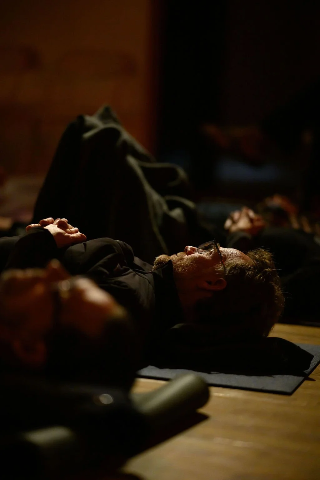 A man with glasses and a beard lying on his back with hands clasped on his chest, in a dark room with other people, meditating and enjoying a sound bath.
