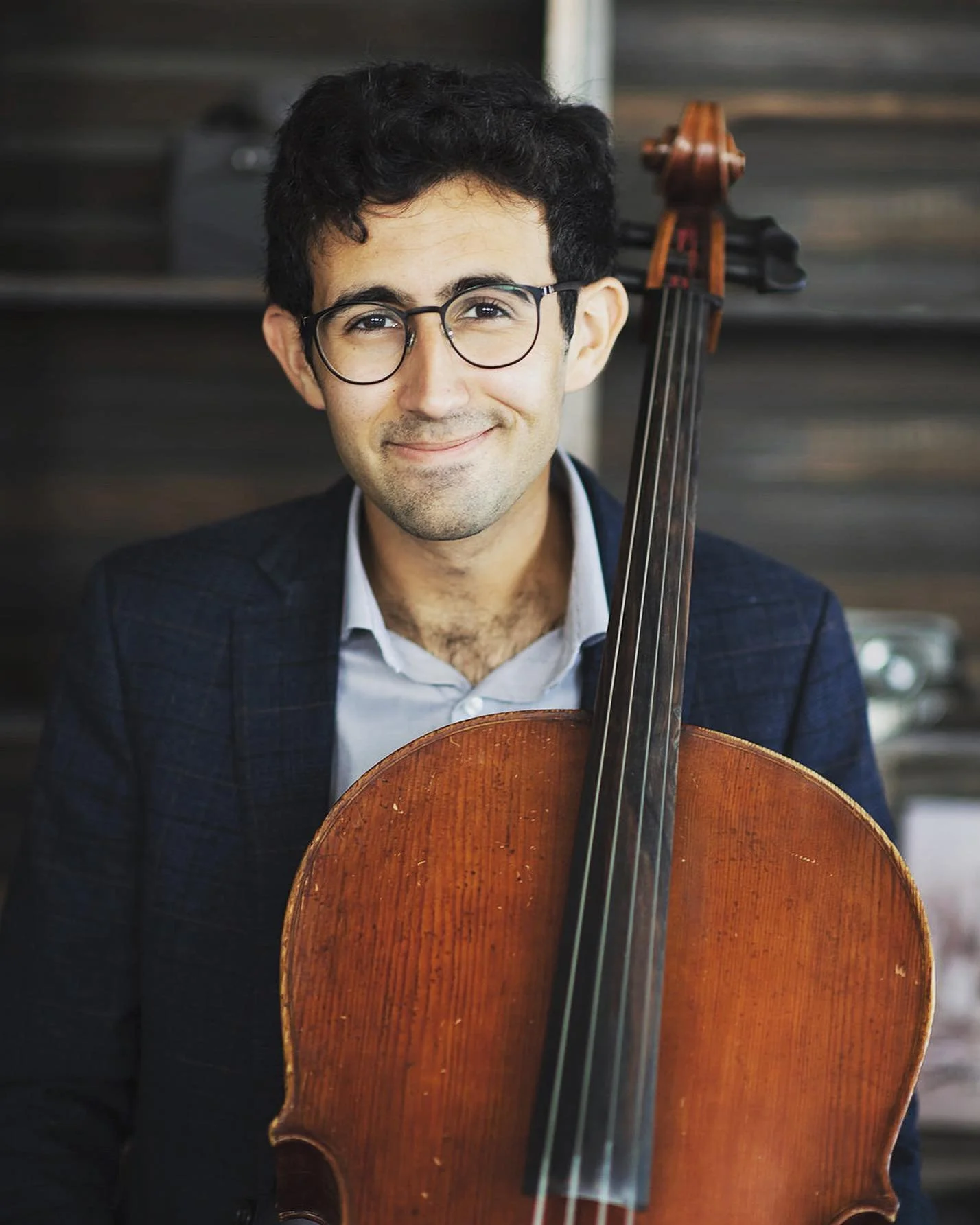 A young man with curly black hair, glasses, and a light beard holding a cello, smiling at the camera against a dark wooden background.