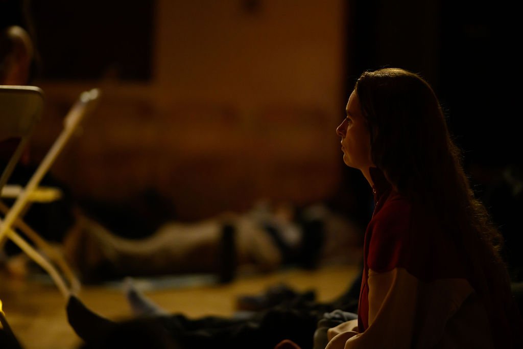 A woman with long dark hair sitting in a dark room, facing to the left, with a serious or contemplative expression. The background is dimly lit and blurred.