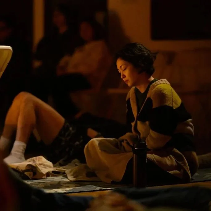 A woman with short dark hair sitting on the floor in a dimly lit room, looking down with a contemplative expression, surrounded by other people in the background.