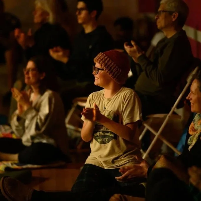 A group of people applauding a performance or presentation, with a young person in the foreground wearing a beige t-shirt and a maroon knit cap, smiling and clapping.