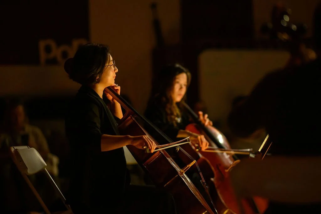 Two women playing cellos in a dimly lit room, during a performance or rehearsal.