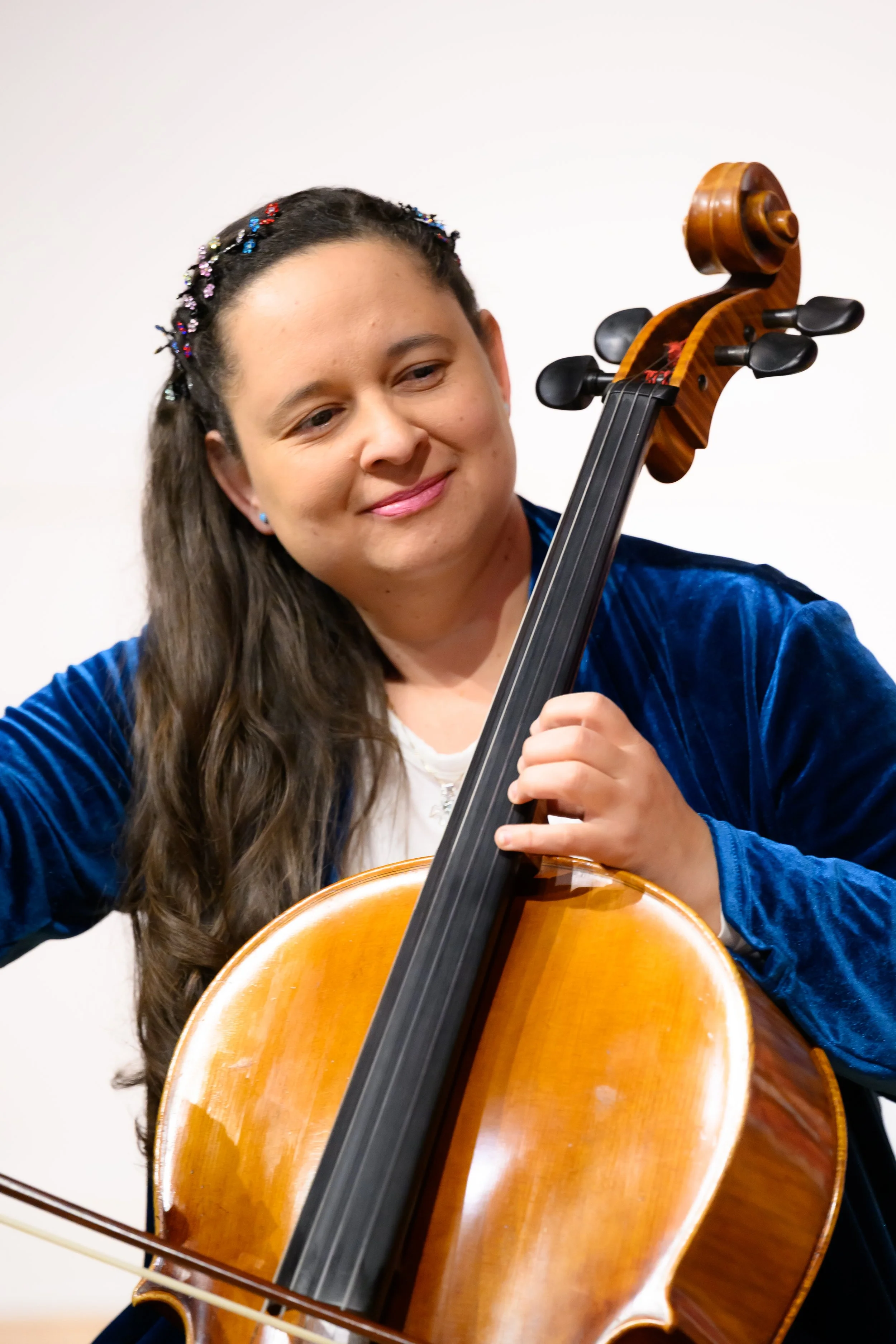 A woman with long brown hair, wearing a blue velvet jacket, playing a cello, with a joyous expression.