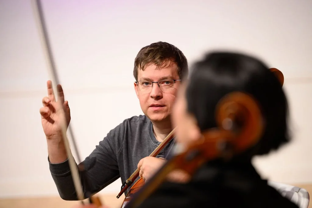 A man with glasses gesturing with his right hand while talking to a woman playing a cello, with a white wall in the background.