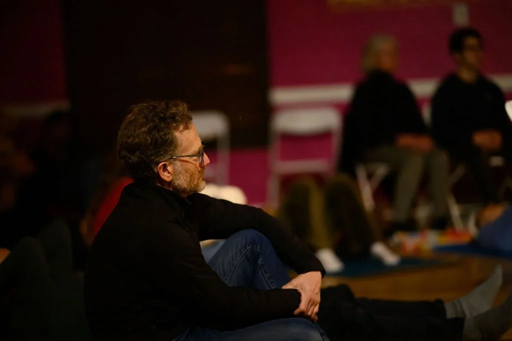 A man with glasses and a beard sitting cross-legged and listening in a dimly lit room, with blurred people sitting on chairs in the background.