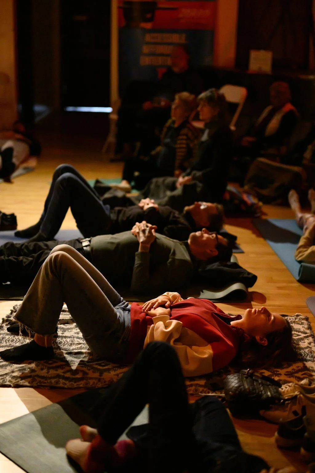 People lying on mats and sitting on chairs in a dimly lit room, participating in a meditation and sound bath session.