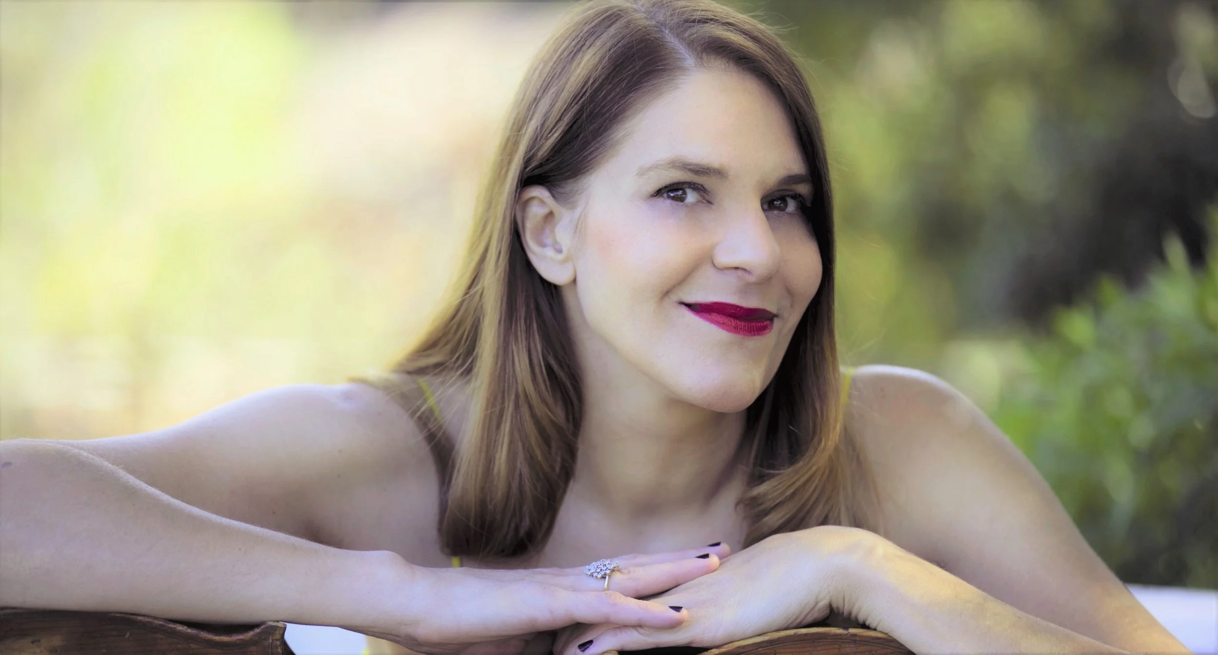 A woman with medium-length brown hair and red lipstick, smiling gently, rests her arms on a cello outdoors with blurred green foliage in the background.
