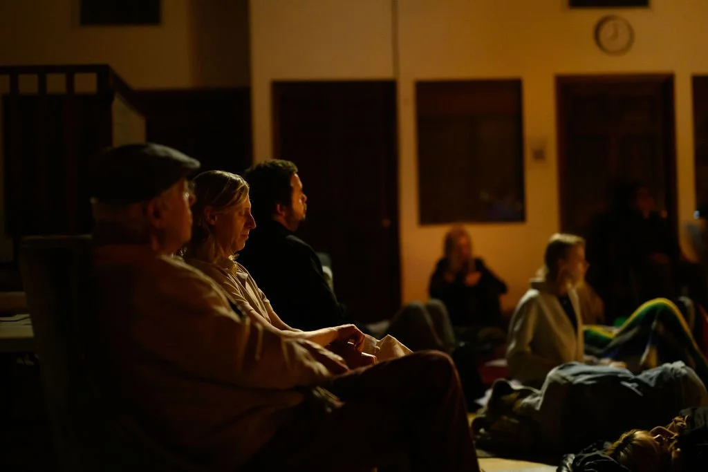 People sitting on chairs and on the floor in a dimly lit room, listening attentively to a live performance.