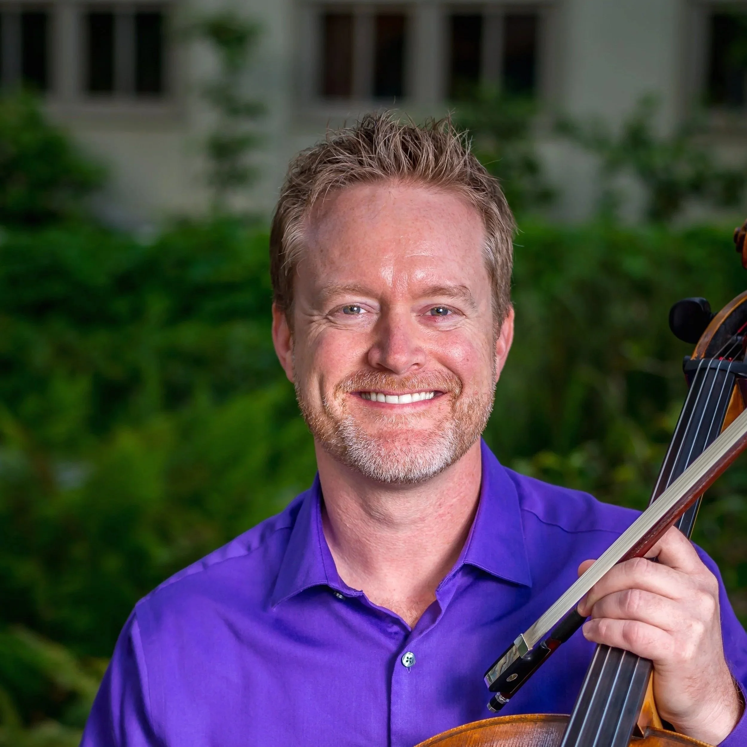 A smiling man with short, light brown hair and a beard, wearing a purple shirt, holding a cello indoors with a green, blurred background.