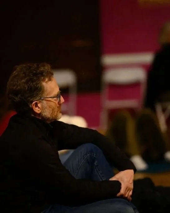 A man with glasses sitting on the floor listening attentively, with a thoughtful expression.