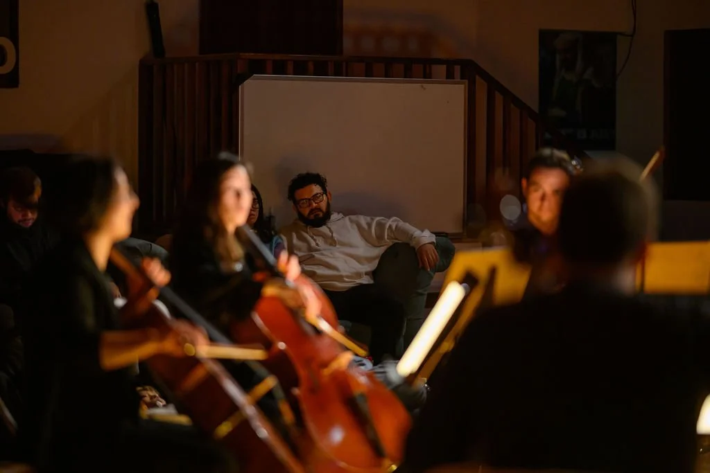 Group of musicians, including a man and women, playing the cello in a dimly lit room, with male audience in the background watching with focused expression.