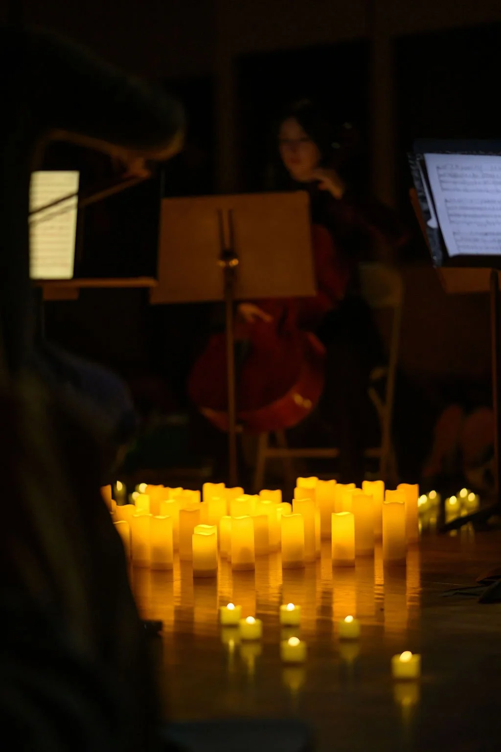 Candles arranged on the floor with a musician playing the cello in the background during an indoor performance with warm ambiance.