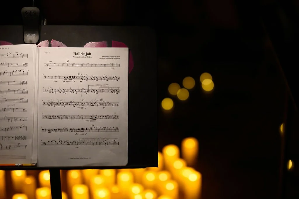 Sheet music titled "Hallelujah" arranged for cello, on a music stand, with blurred candle lights in the background.