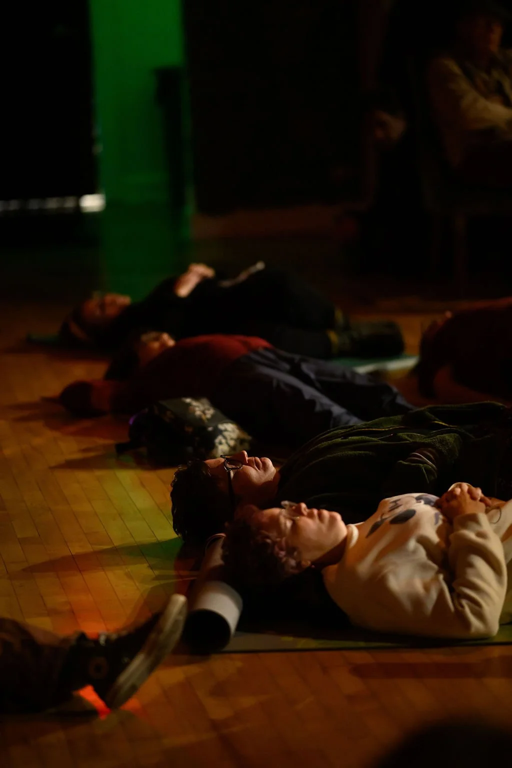 People lying on the floor in a dark room, illuminated by dim, colorful lighting, participating in a meditation, sound bath, or relaxation activity.