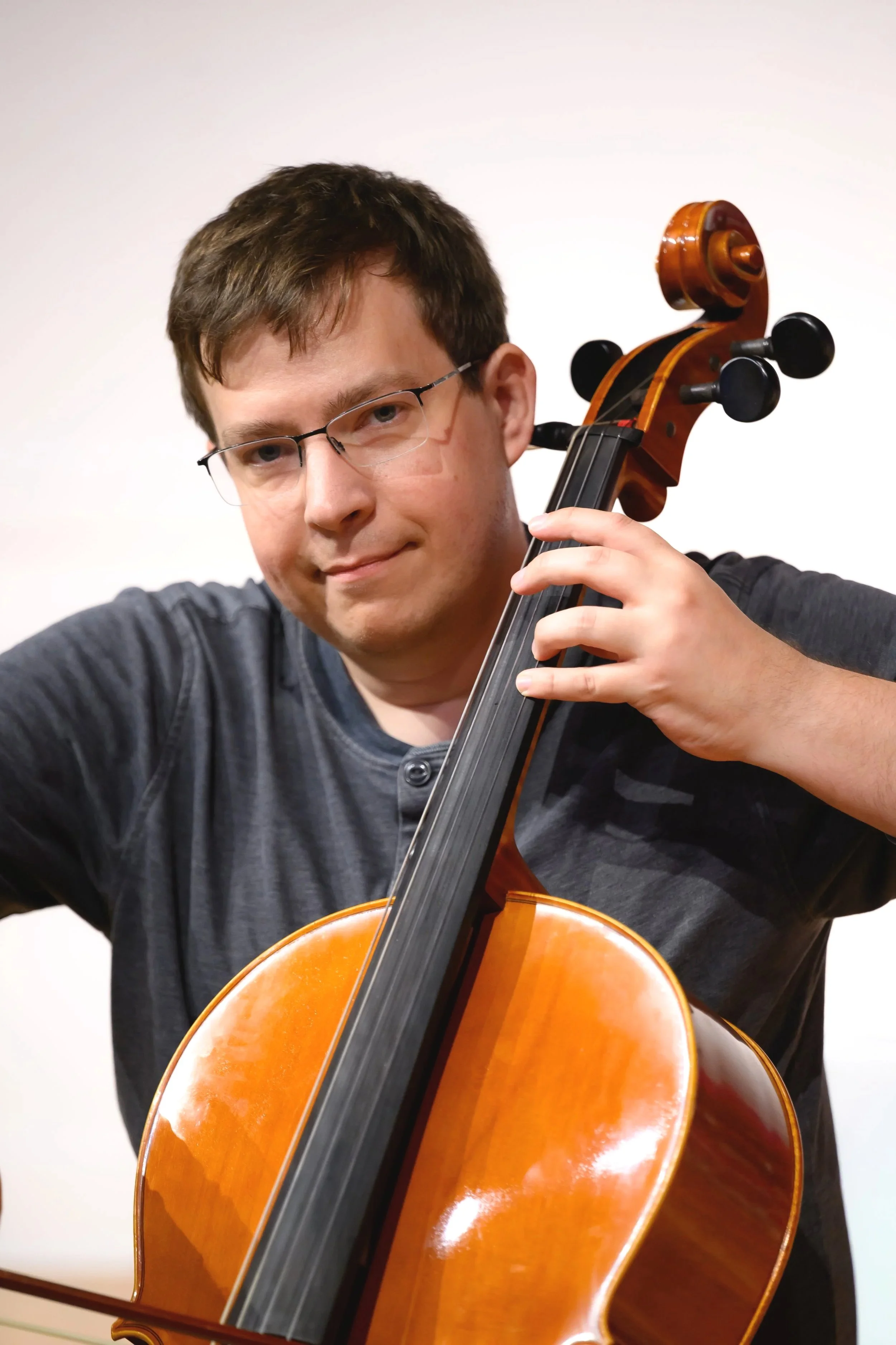 A young man with glasses playing a cello with a smile.