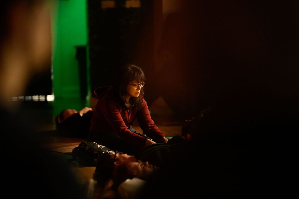 A woman with glasses in a dark room, stretching her legs on the floor with a reflective expression, illuminated by dim lighting