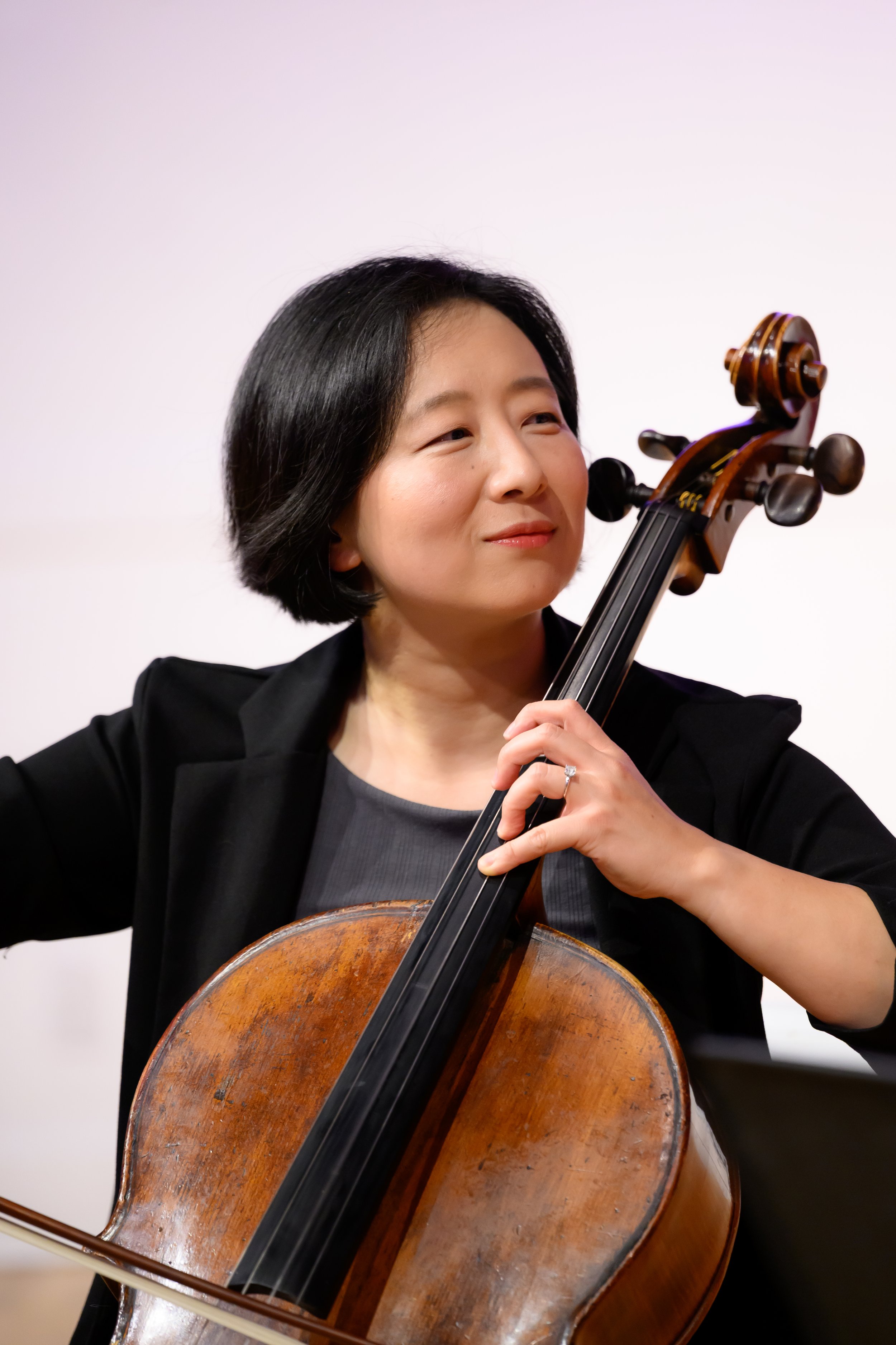 A woman in short black hair playing a cello, dressed in black, with a focused expression, against a plain light background.