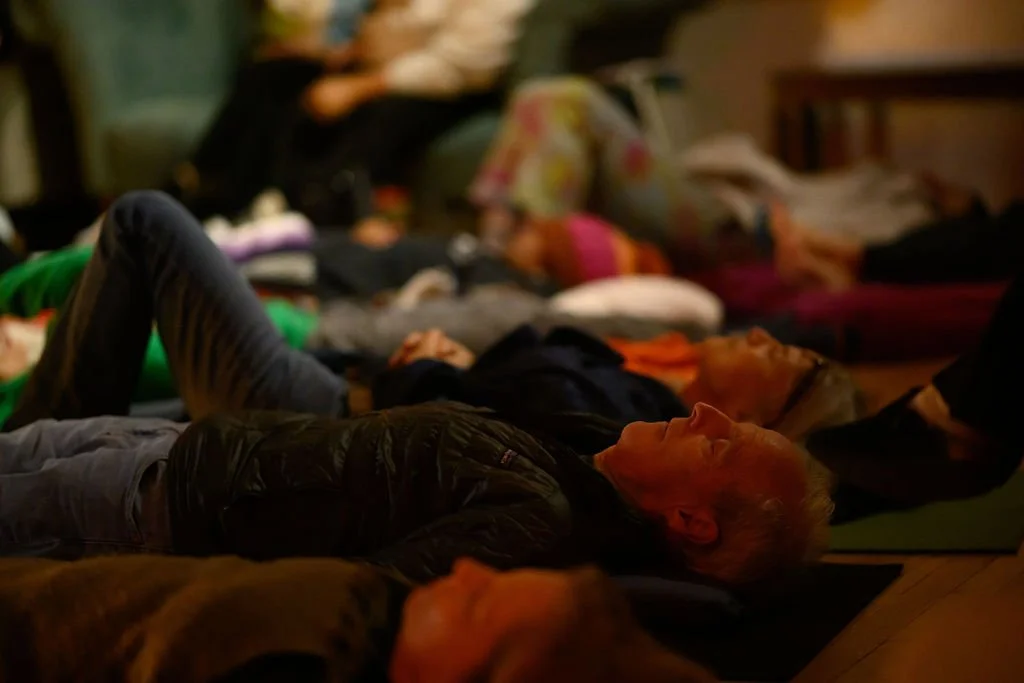 People lying on yoga mats on the floor in a dimly lit room, possibly during a meditation, sound bath, or relaxation session.