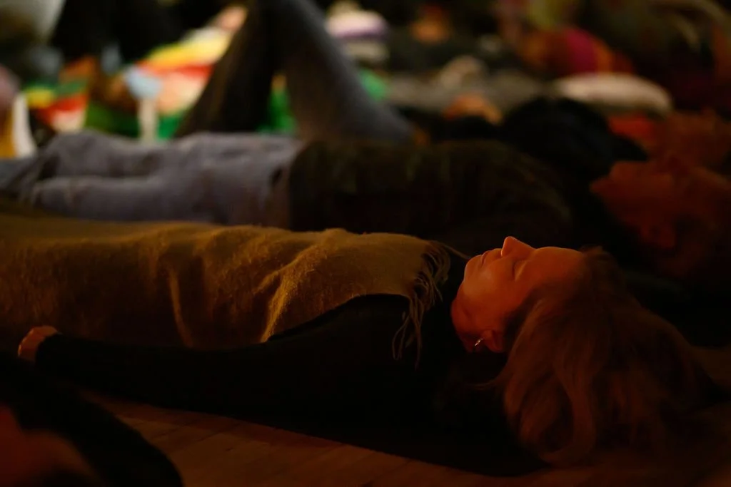 A woman lying down on her back with closed eyes, surrounded by other people lying down in a dimly lit setting, participating in a sound bath.