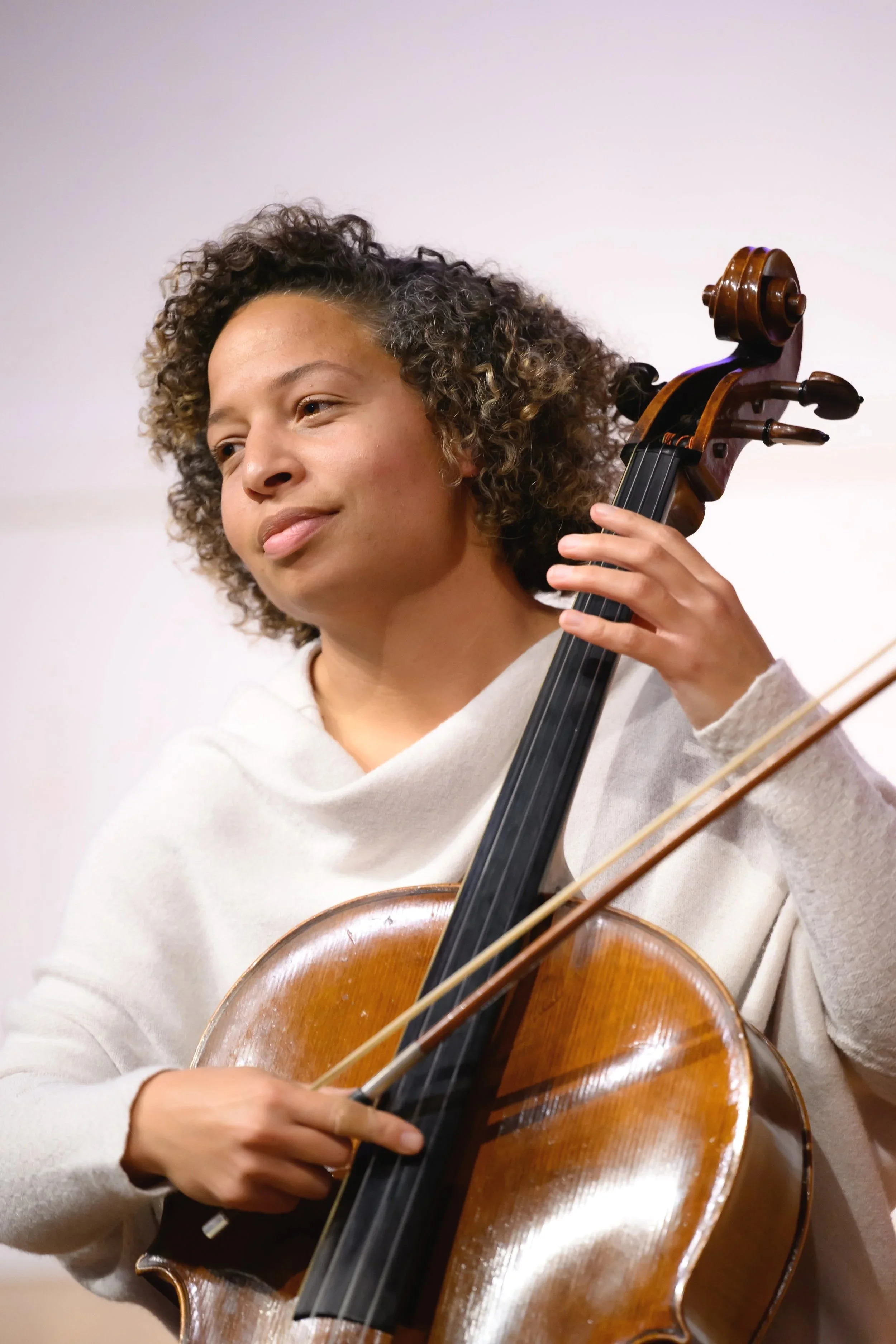 A woman with curly hair playing a cello, with a smile on her face.