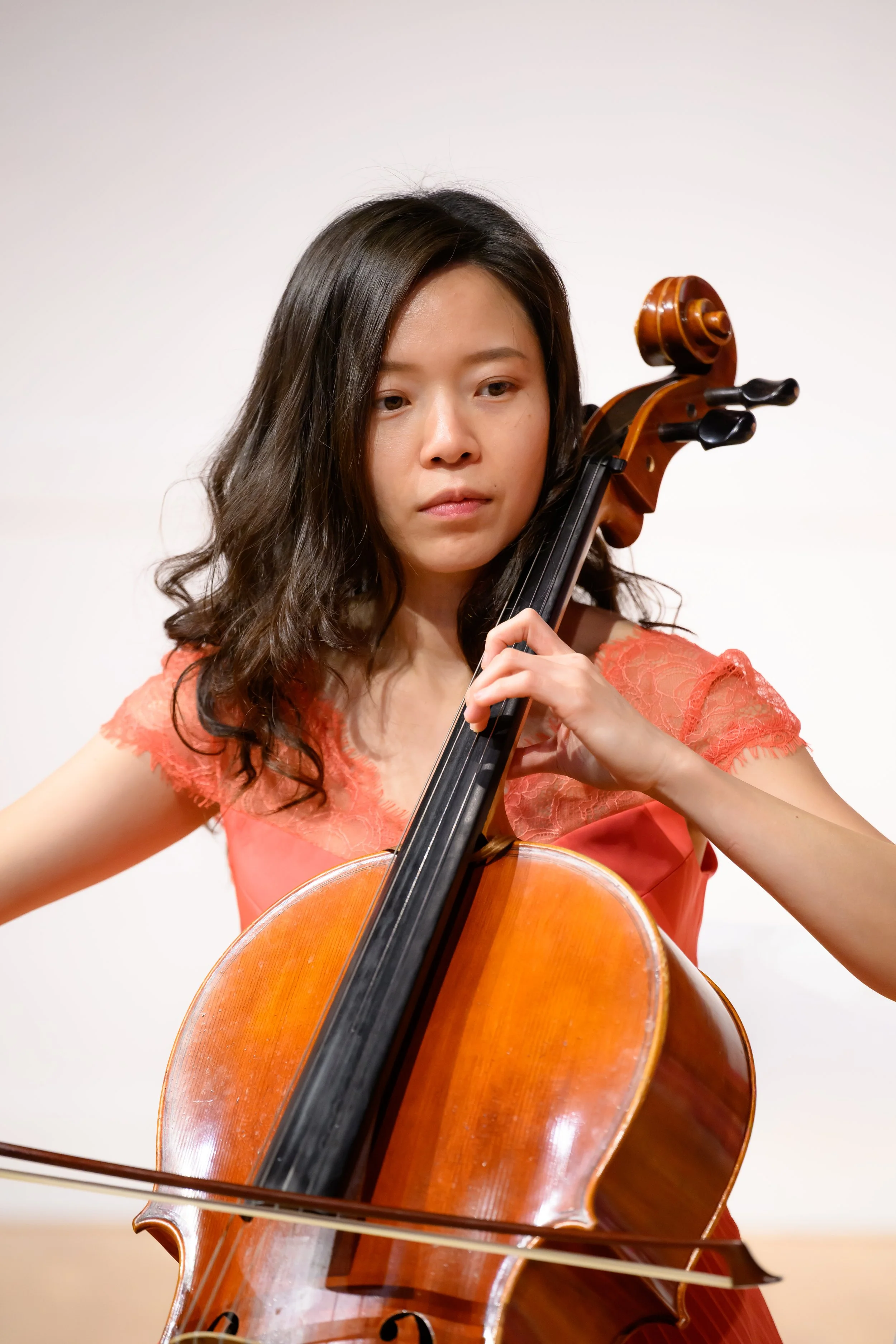 A young woman playing a cello, wearing a coral dress with lace sleeves, with a focused expression.