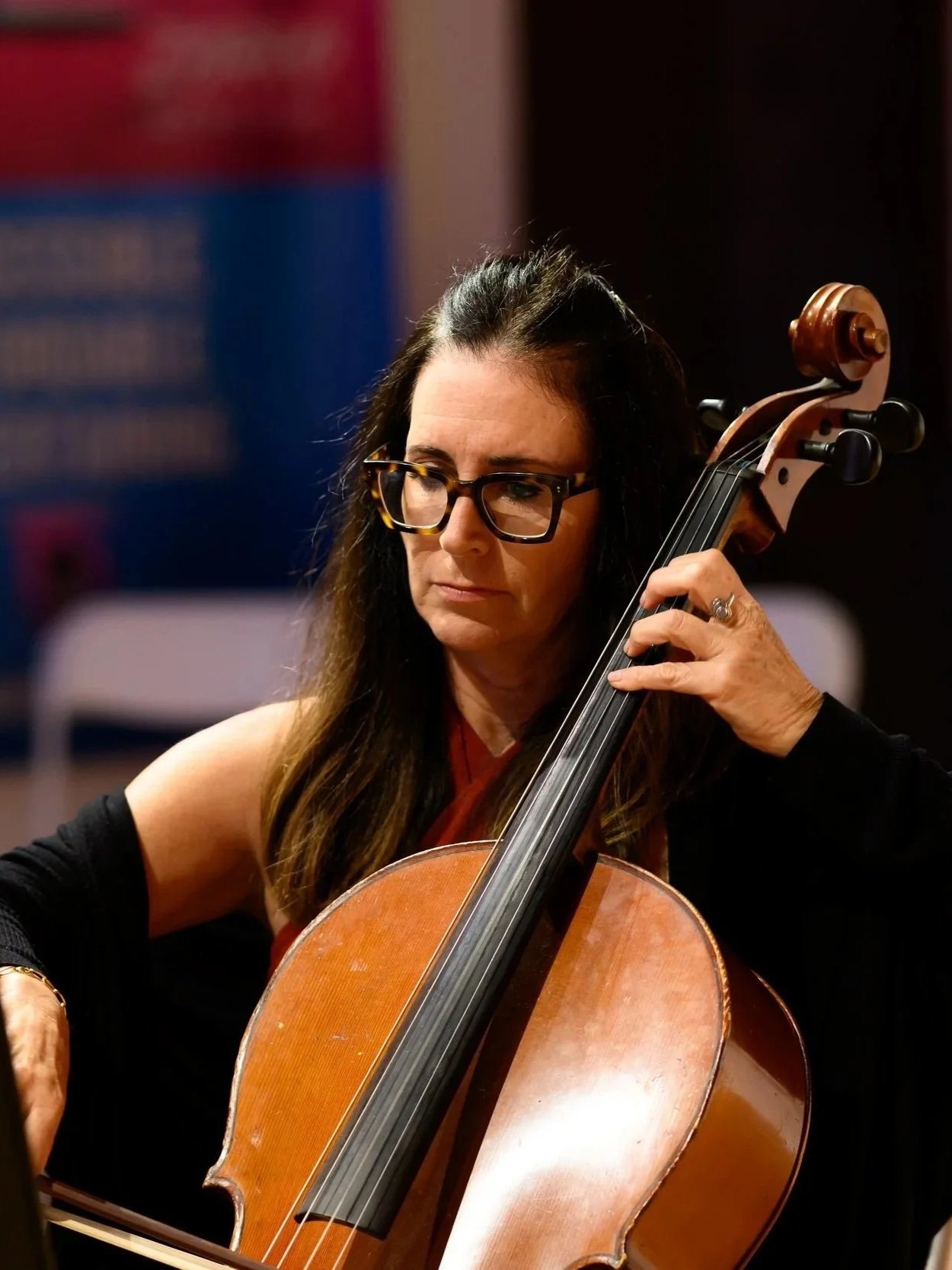 A woman with dark long hair, glasses, and a black outfit playing a cello, with a focused expression.
