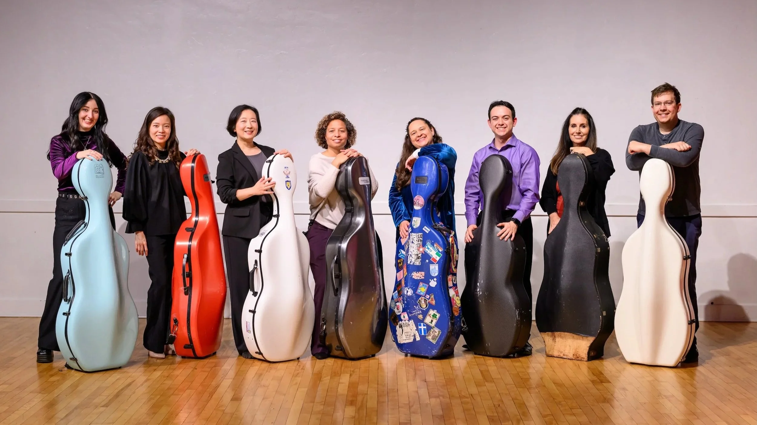 A group of culturally diverse musicians standing indoors, each holding a different colored cello case, with wooden flooring and plain wall background.