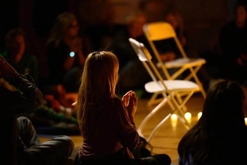A young woman is kneeling with her hands clasped in front of her, participating in a sound bath meditation session in a dimly lit room, with other meditators in the background.
