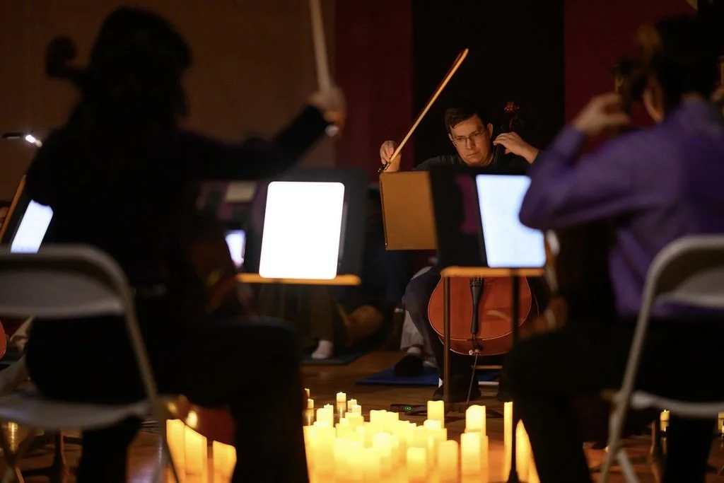 An orchestra rehearsal with musicians playing string instruments in a dimly lit room, illuminated by warm candlelight on the floor.