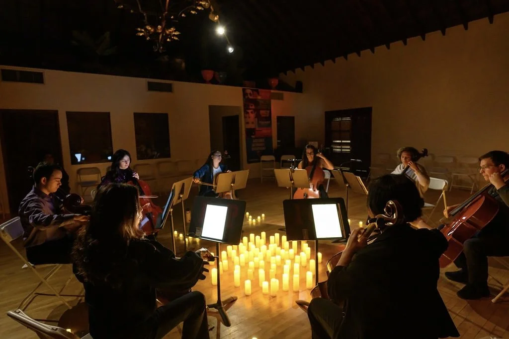 A group of cellists performing in a dimly lit room with candles arranged in the shape of a heart on the floor.
