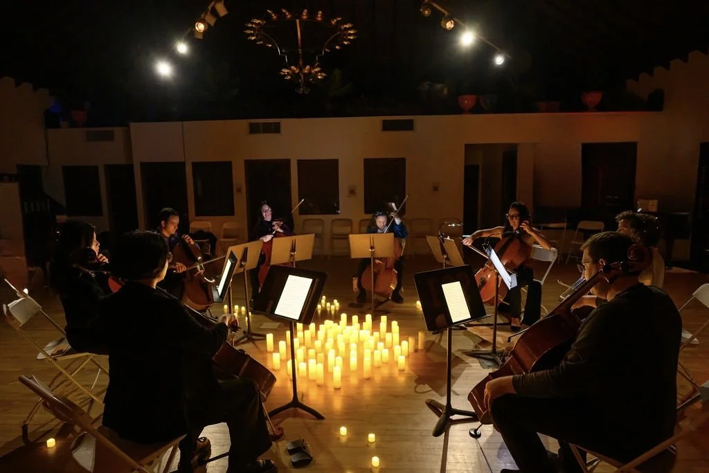 A group of musicians playing cellos during a candlelit rehearsal or performance in a room lit by candles.