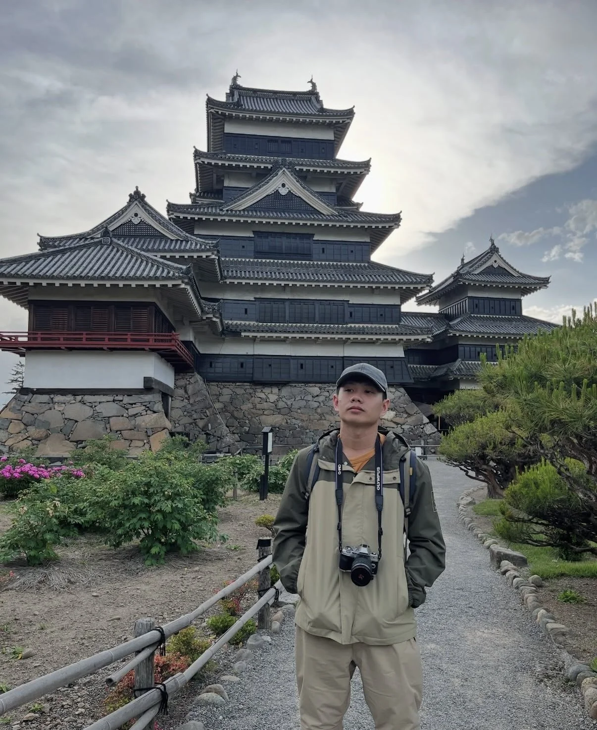 Joven con mochila y cámara en un parque frente a un castillo tradicional japonés con techo de teja y paredes blancas, bajo cielo nublado.