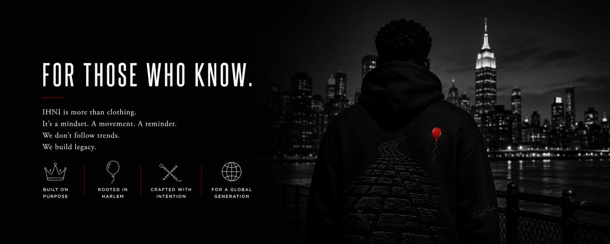 A person wearing a black hoodie with a red balloon graphic on the back standing on a waterfront with a city skyline at night, including the Empire State Building.
