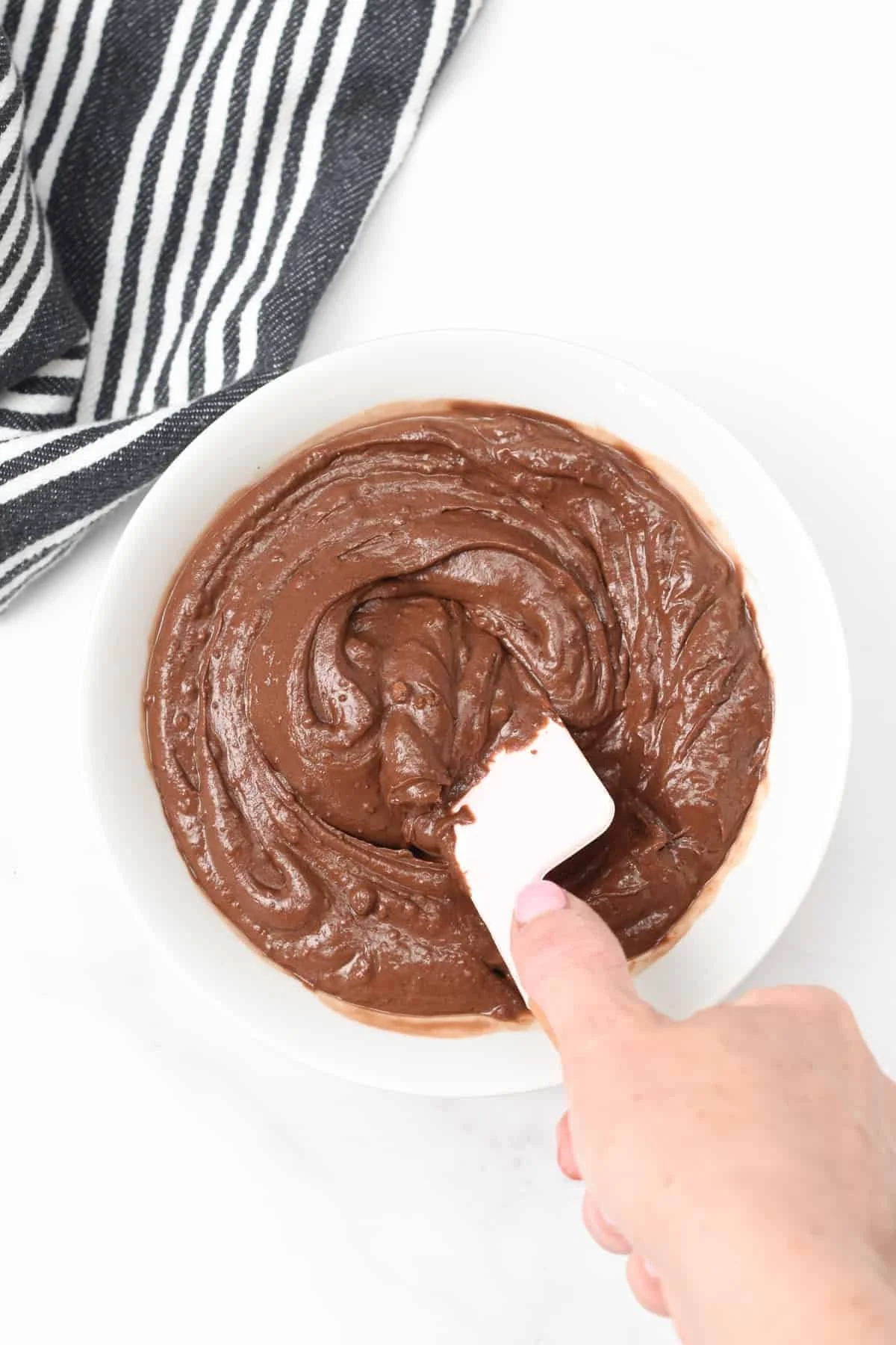 A hand using a spatula to spread chocolate frosting in a circular motion in a white bowl on a white surface, with a black and white striped towel nearby.