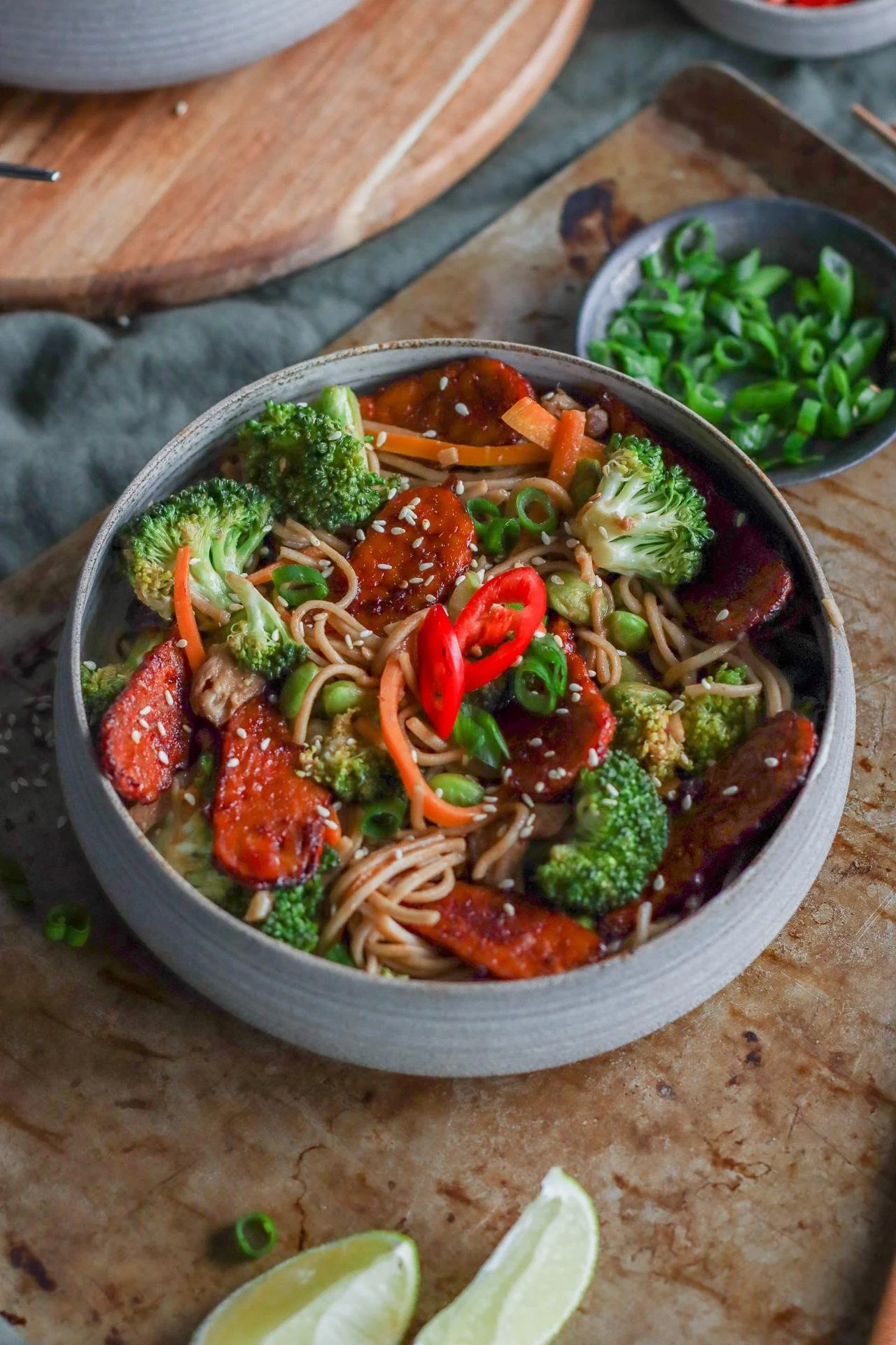 A bowl of Asian vegetable stir-fry with broccoli, carrots, and red peppers on noodles, garnished with sesame seeds and sliced green onions, on a rustic wooden surface with lime wedges nearby.
