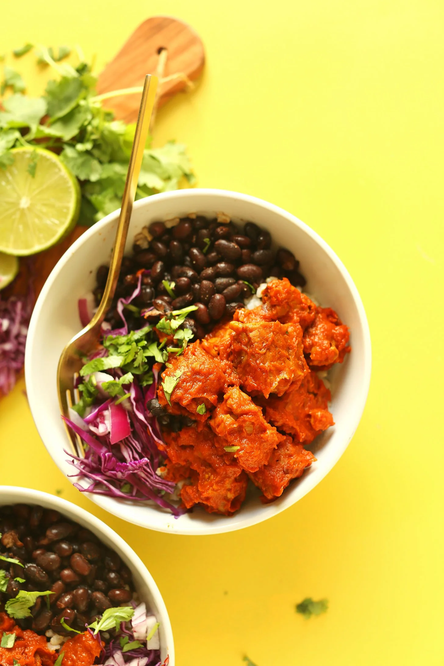 Bowl of red chili with black beans, garnished with cilantro, served with shredded purple cabbage. Background includes lime slices, green leafy herbs, and a wooden cutting board on a yellow surface.