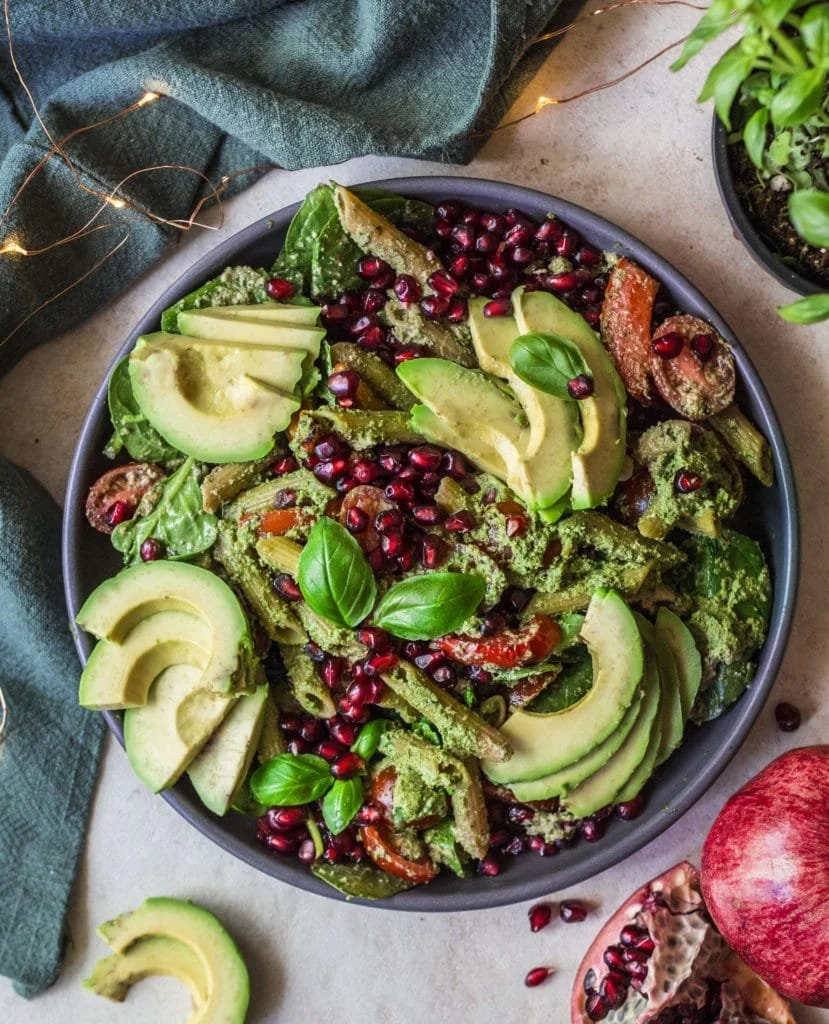 A bowl of salad containing sliced avocados, green vegetables, pomegranate seeds, and basil leaves, surrounded by additional ingredients like an apple and a pomegranate, with decorative string lights and a green cloth nearby.