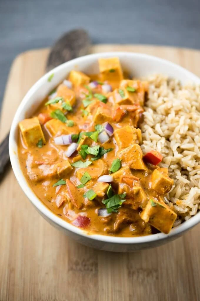 A bowl of Indian curry with rice, garnished with chopped cilantro and diced red onions, served on a wooden board.