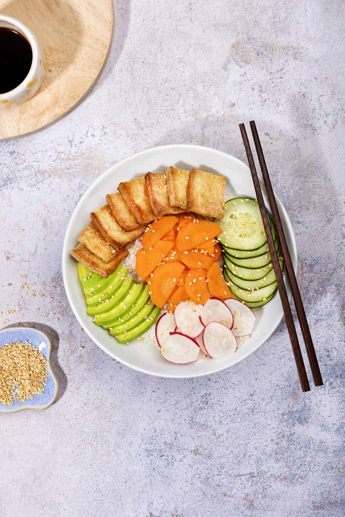 A bowl of fresh vegetable salad with sliced cucumbers, radishes, shredded carrots, and lettuce, topped with sesame seeds, with fried tofu pieces on the side, chopsticks resting on the bowl, and a glass of soy sauce on a wooden tray nearby.