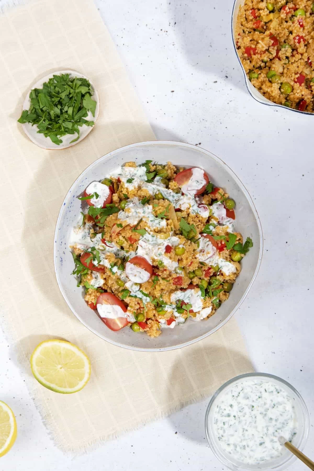 A bowl of couscous salad with cherry tomato slices, peas, and fresh herbs, topped with a creamy white dressing. Surrounding it are lemon halves, chopped parsley, and a small bowl of herb dressing on a white surface with a beige cloth.