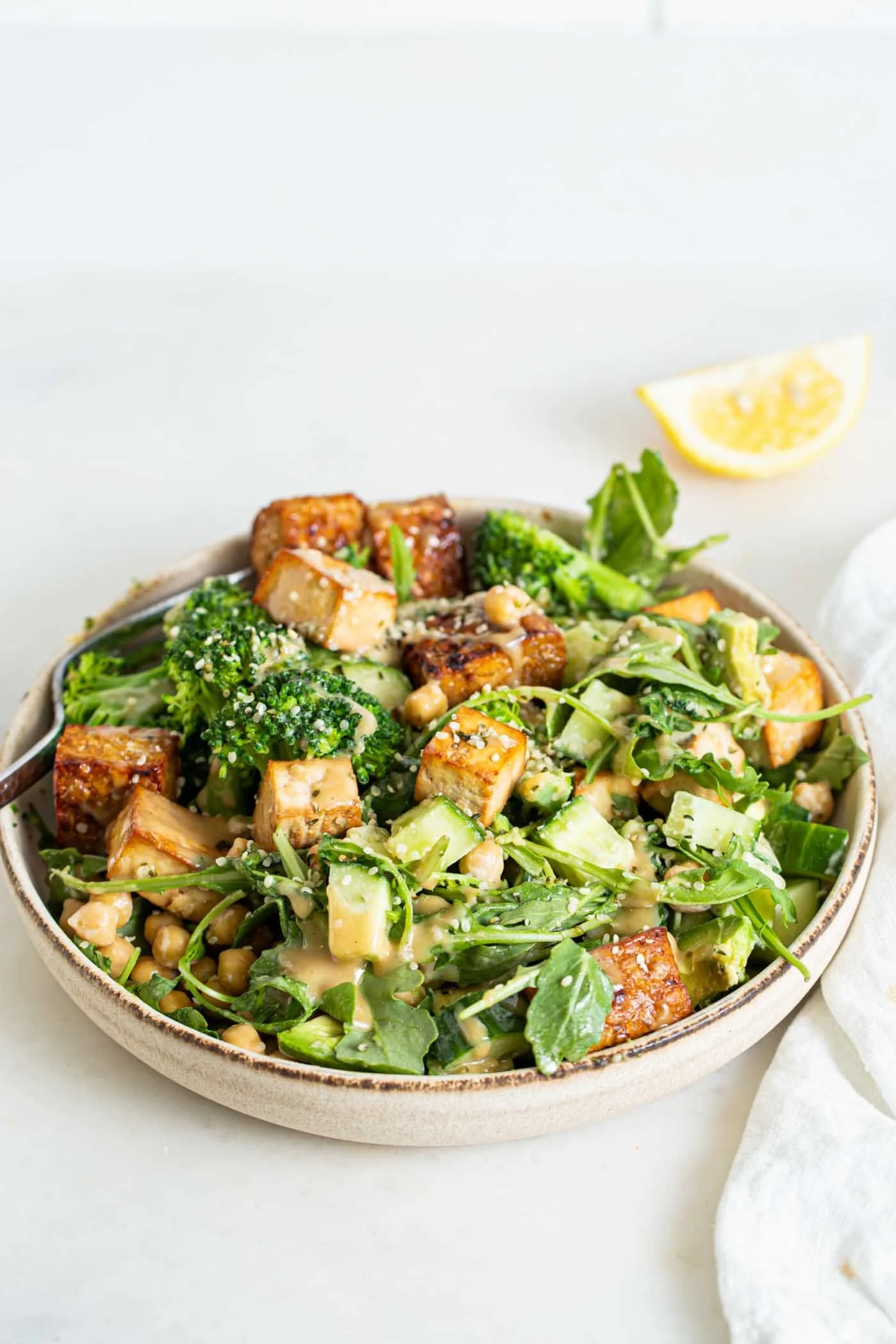 A bowl of salad with tofu, broccoli, chickpeas, cucumbers, and greens, garnished with Parmesan cheese and served with a lemon wedge.
